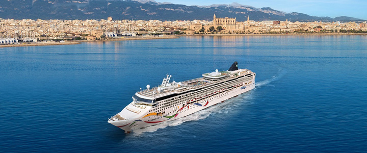 NCL Kreuzfahrtschiff umgeben von ruhigem Gewässer; Gebäude im Hintergrund.